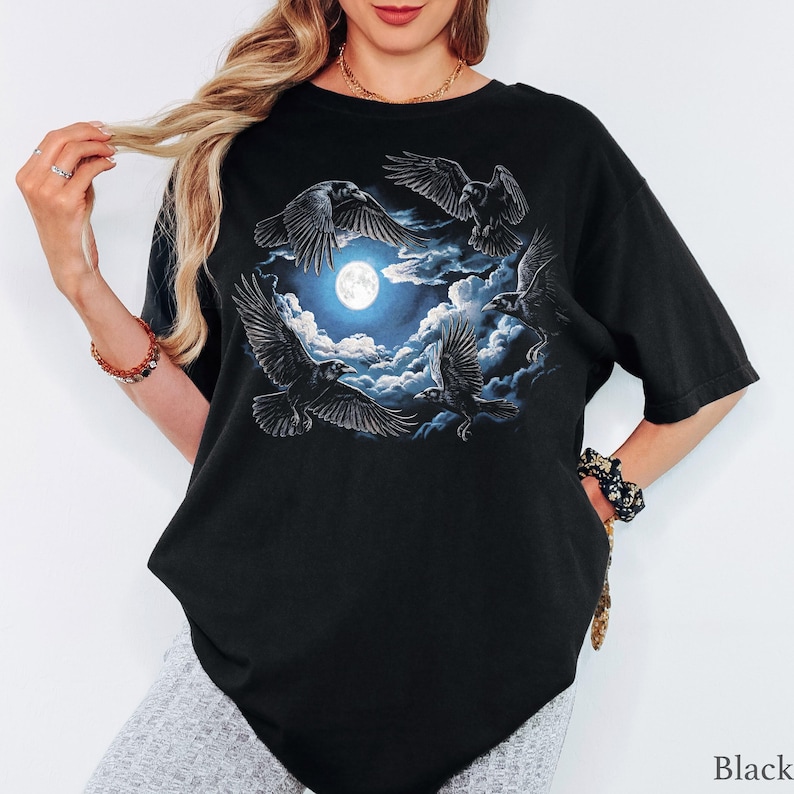 May include: A black short-sleeved t-shirt featuring a graphic print of four black ravens flying in a circular formation around a bright, full moon set against a backdrop of dark, swirling blue clouds. The design is centered on the chest area.