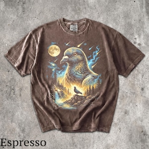 May include: A brown short-sleeve t-shirt featuring a graphic print of a large pigeon head above a smaller pigeon perched on a rock near a waterfall, surrounded by a forest, a full moon, and falling stars with blue light streaks in the background.