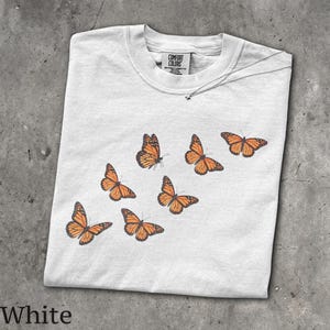 May include: A white short-sleeve t-shirt featuring a graphic print of six orange and black monarch butterflies arranged in a diagonal pattern. The inner neck label displays the text Comfort Colors, indicating the brand of the garment.