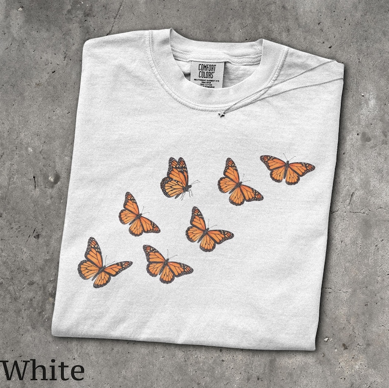 May include: A white short-sleeve t-shirt featuring a graphic print of six orange and black monarch butterflies arranged in a diagonal pattern. The inner neck label displays the text Comfort Colors, indicating the brand of the garment.
