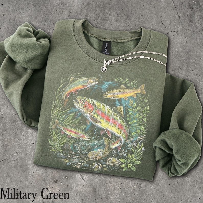 May include: A military green crewneck sweatshirt featuring a graphic print of three trout swimming among aquatic plants and rocks. The design includes a large central fish with a red stripe, a smaller fish above, and one below, all rendered in natural colors.