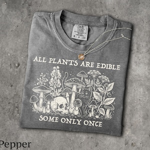 May include: A grey t-shirt featuring a white graphic print of mushrooms, a skull, and a cat. The text reads ALL PLANTS ARE EDIBLE SOME ONLY ONCE. A thin gold-toned necklace with a small circular pendant rests on the neckline of the shirt.