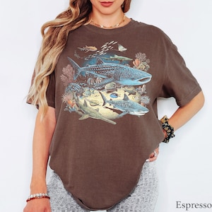 May include: An espresso-colored short-sleeve t-shirt featuring a detailed graphic print of various marine life, including a large whale shark, a tiger shark, a sawfish, and smaller fish swimming among coral reef elements in shades of blue and tan.