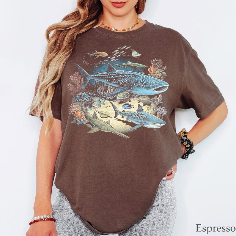 May include: An espresso-colored short-sleeve t-shirt featuring a detailed graphic print of various marine life, including a large whale shark, a tiger shark, a sawfish, and smaller fish swimming among coral reef elements in shades of blue and tan.