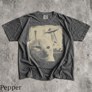 May include: A charcoal grey short-sleeve t-shirt featuring a graphic print of a cat with two antennae on its head. Above the cat, a flying saucer beams down a cow toward a farm building. The design has a faded, vintage-inspired aesthetic.