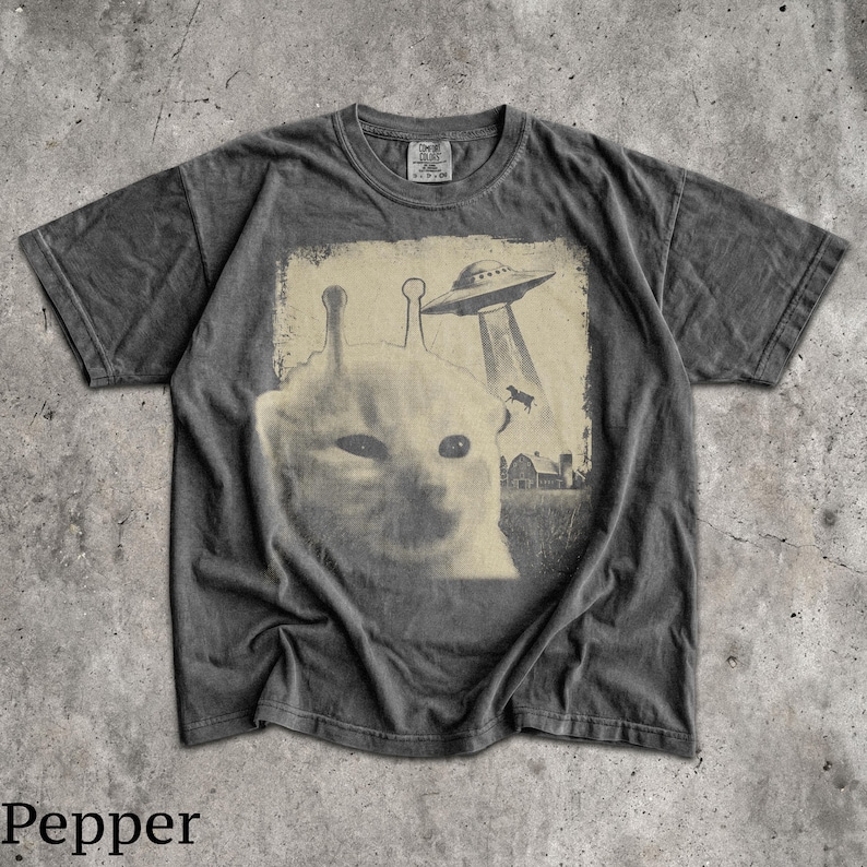 May include: A charcoal grey short-sleeve t-shirt featuring a graphic print of a cat with two antennae on its head. Above the cat, a flying saucer beams down a cow toward a farm building. The design has a faded, vintage-inspired aesthetic.