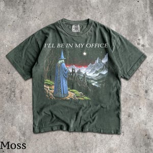 Op de afbeelding: Een donkergroen T-shirt met korte mouwen en een grafische print van een tovenaar in een blauwe mantel die op een klif staat en uitkijkt over een berglandschap onder een sterrenhemel. De tekst I'LL BE IN MY OFFICE staat in wit erboven.