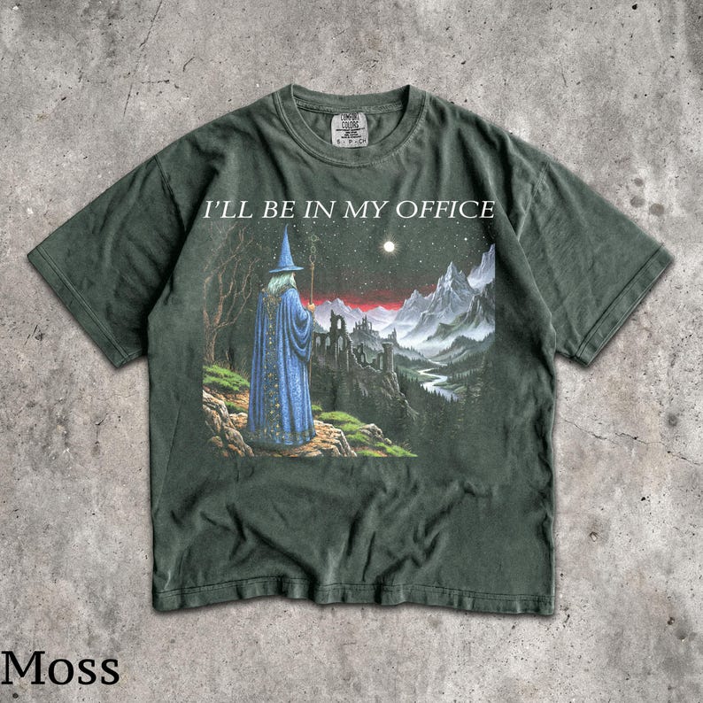 Op de afbeelding: Een donkergroen T-shirt met korte mouwen en een grafische print van een tovenaar in een blauwe mantel die op een klif staat en uitkijkt over een berglandschap onder een sterrenhemel. De tekst I'LL BE IN MY OFFICE staat in wit erboven.