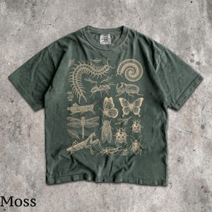 May include: A dark moss green short-sleeve t-shirt featuring a detailed, light-colored graphic print of various insects, including a centipede, butterfly, dragonfly, and beetles, arranged in a grid-like pattern across the front of the garment.