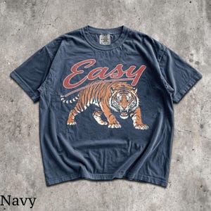May include: A navy blue short-sleeve t-shirt featuring a graphic of a prowling tiger in orange, black, and white. Above the tiger, the word "Easy" is printed in a large, stylized red font. The shirt has a crew neckline and a visible brand tag.