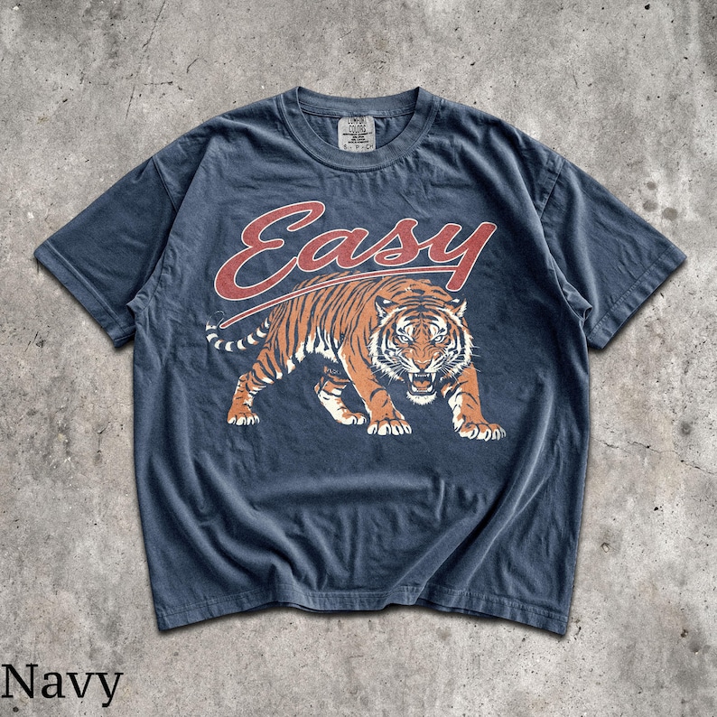 May include: A navy blue short-sleeve t-shirt featuring a graphic of a prowling tiger in orange, black, and white. Above the tiger, the word "Easy" is printed in a large, stylized red font. The shirt has a crew neckline and a visible brand tag.