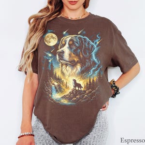 May include: A brown short-sleeved t-shirt featuring a graphic print of a Bernese Mountain Dog head above a smaller dog standing on a rocky cliff near a waterfall and forest, with a full moon and shooting stars in a blue and yellow night sky.