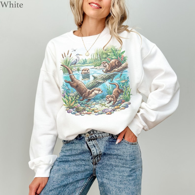 Op de afbeelding: Een wit sweatshirt met lange mouwen en een kleurrijke grafische print van otters die in een rivier zwemmen tussen waterplanten, rotsen en een reiger. Het ontwerp toont een gedetailleerd onderwatersc&egrave;ne met kiezels en diverse waterplanten.
