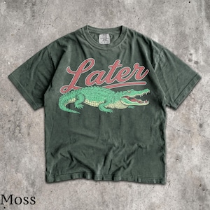 Puede incluir: Camiseta verde musgo con un dise&ntilde;o de cocodrilo verde y la palabra "Later" en letras rojas. La camiseta tiene un aspecto vintage. La palabra "Moss" est&aacute; impresa en la parte inferior de la imagen.