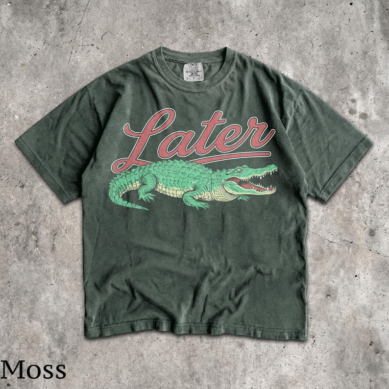 Puede incluir: Camiseta verde musgo con un dise&ntilde;o de cocodrilo verde y la palabra "Later" en letras rojas. La camiseta tiene un aspecto vintage. La palabra "Moss" est&aacute; impresa en la parte inferior de la imagen.