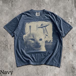 May include: A navy blue short-sleeve t-shirt featuring a graphic print of a cat with two antennae on its head. Above the cat, a flying saucer beams down a cow toward a farm building. The print has a distressed, vintage-inspired aesthetic.