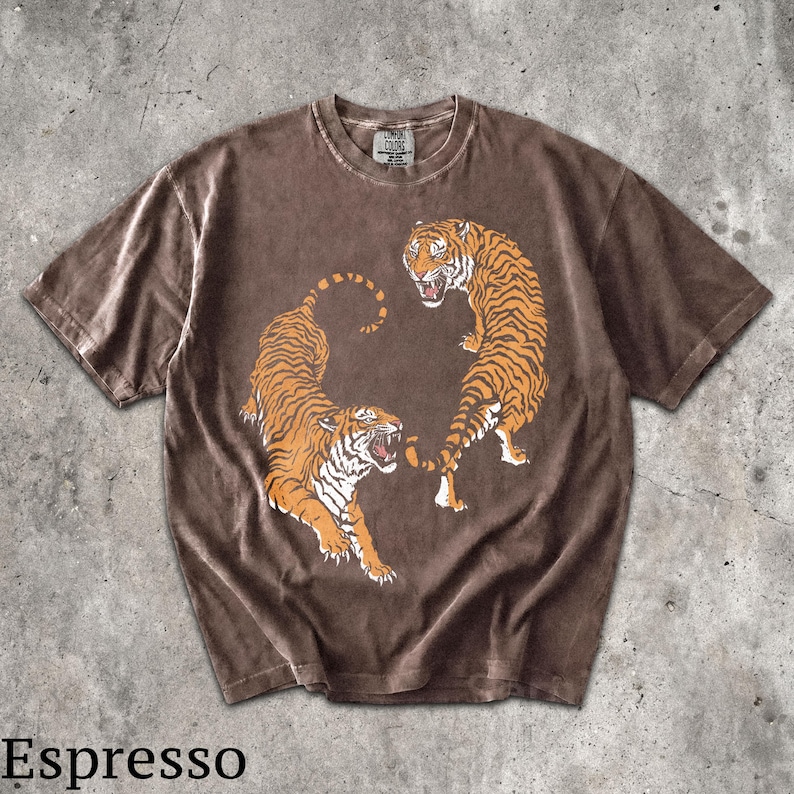 May include: A short-sleeve espresso brown t-shirt featuring a graphic of two orange and black tigers in a dynamic pose. The shirt has a crew neckline and a Comfort Colors brand label printed on the inside of the collar. The fabric has a washed look.