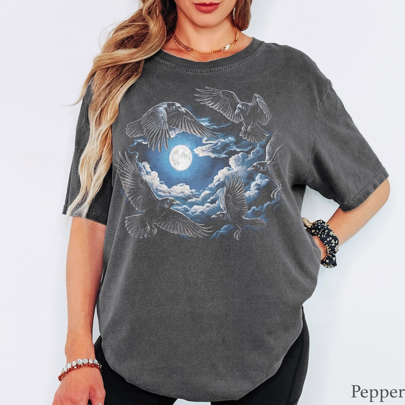 May include: A charcoal gray short-sleeve t-shirt featuring a graphic print of four black ravens flying around a bright full moon surrounded by blue clouds. The shirt has a crew neckline and a relaxed fit with a faded, vintage-style wash finish.