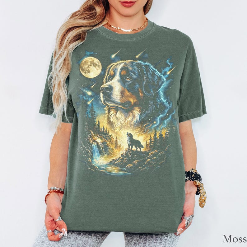 May include: A moss green short-sleeve t-shirt featuring a graphic print of a Bernese Mountain Dog portrait above a smaller silhouette of a dog standing on a rocky cliff near a waterfall, a full moon, and falling stars in the night sky.