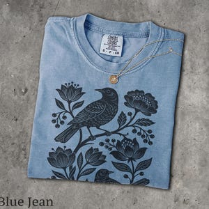 May include: A blue jean colored t-shirt featuring a black graphic print of two birds perched among flowers and berries. The shirt has a crew neckline with a Comfort Colors brand label showing size S, and a gold-toned necklace rests on the collar.