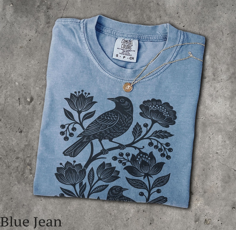 May include: A blue jean colored t-shirt featuring a black graphic print of two birds perched among flowers and berries. The shirt has a crew neckline with a Comfort Colors brand label showing size S, and a gold-toned necklace rests on the collar.