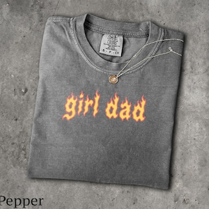 May include: A pepper-colored short-sleeve t-shirt with a crew neckline. The chest features the text 'girl dad' printed in a stylized orange and yellow flame font. A thin silver-toned chain necklace with a small circular pendant rests on the collar.