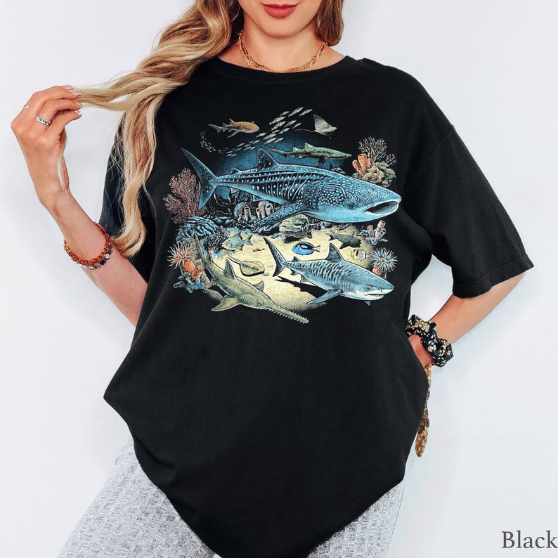 May include: A black short-sleeve t-shirt featuring a detailed graphic print of various marine life, including a large whale shark, a tiger shark, a sawfish, and smaller fish swimming among coral reefs and underwater rock formations.