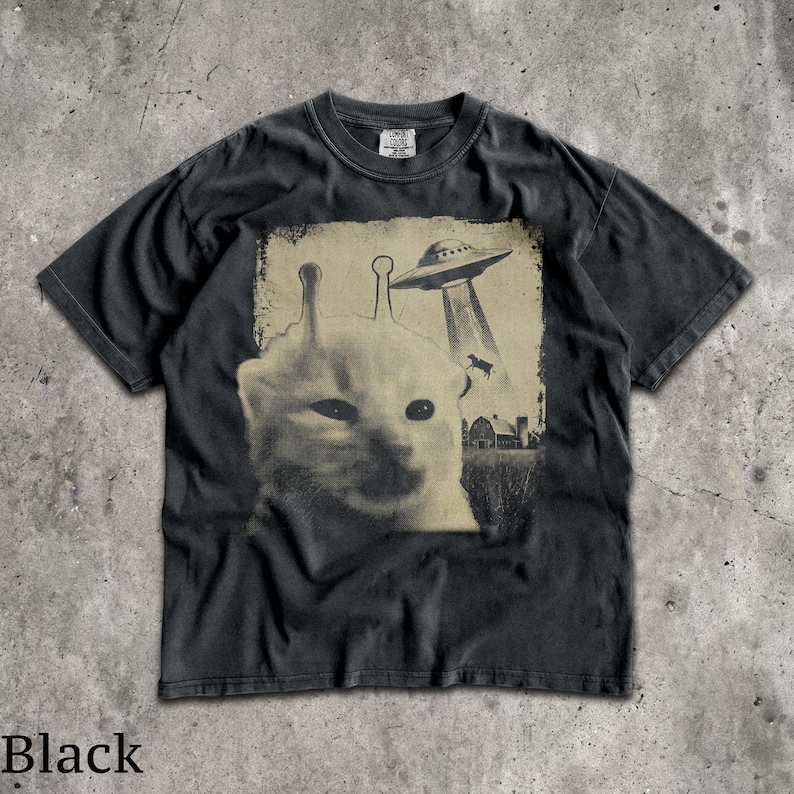 May include: A black short-sleeve t-shirt featuring a graphic print of a cat with two antennae on its head. Above the cat, a flying saucer hovers over a farm scene with a cow being abducted. The shirt has a crew neckline and a vintage-style wash.