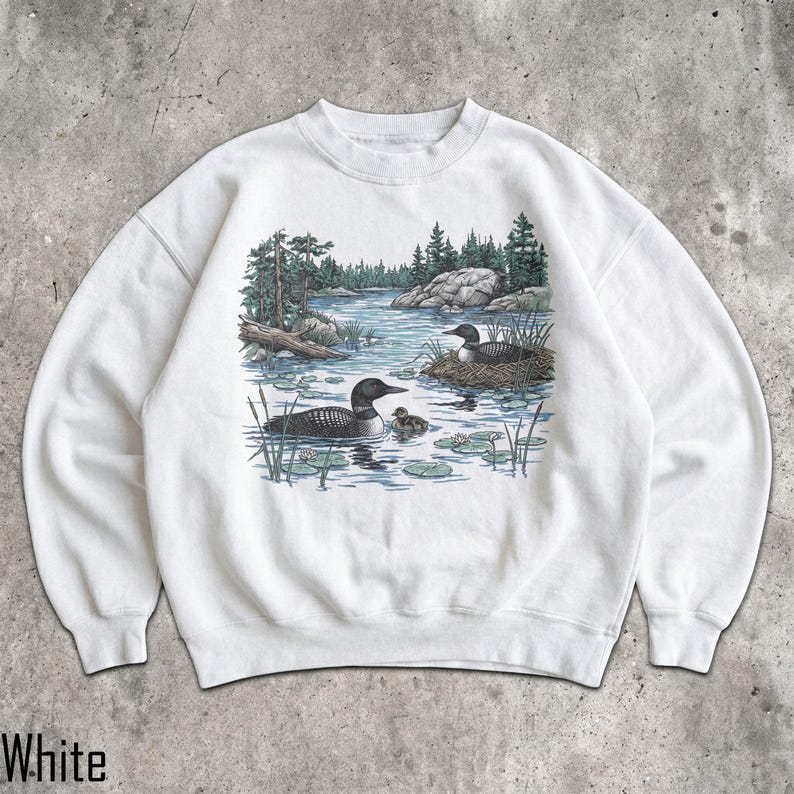 May include: A white crewneck sweatshirt featuring a detailed graphic print of two loons swimming in a lake with a small chick nearby. The scene includes a nest on the water, green pine trees, rocky shorelines, and lily pads in blue and green tones.