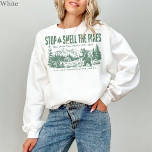 Pode incluir: Uma camisola branca de manga comprida com uma impress&atilde;o gr&aacute;fica verde de um urso a caminhar numa floresta com montanhas e uma cabana. O texto diz STOP AND SMELL THE PINES, Three Billion Trees Planted 1933-1942 e CIVILIAN CONSERVATION CORPS.