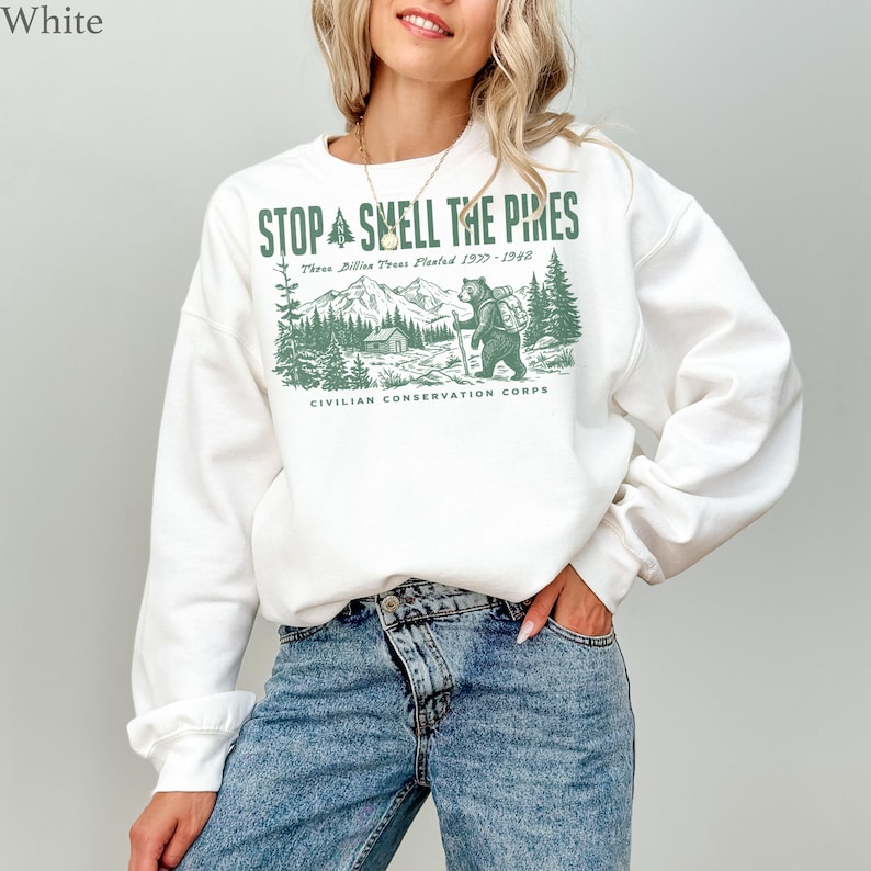 Pode incluir: Uma camisola branca de manga comprida com uma impress&atilde;o gr&aacute;fica verde de um urso a caminhar numa floresta com montanhas e uma cabana. O texto diz STOP AND SMELL THE PINES, Three Billion Trees Planted 1933-1942 e CIVILIAN CONSERVATION CORPS.