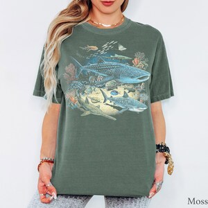 May include: A moss green short-sleeve t-shirt featuring a detailed graphic print of various marine life, including a large whale shark, a tiger shark, and a sawfish swimming among colorful coral reefs and smaller fish in shades of blue and orange.