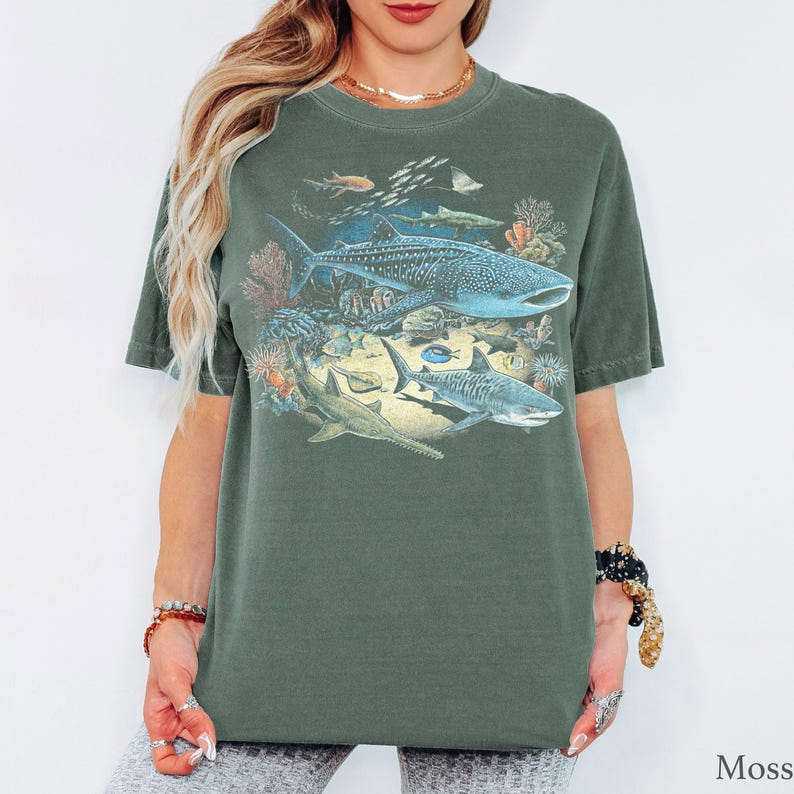 May include: A moss green short-sleeve t-shirt featuring a detailed graphic print of various marine life, including a large whale shark, a tiger shark, and a sawfish swimming among colorful coral reefs and smaller fish in shades of blue and orange.