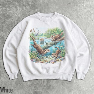 Op de afbeelding: Een wit sweatshirt met ronde hals en een kleurrijke grafische print van otters die in een rivier zwemmen. De illustratie toont meerdere otters, een reiger, waterplanten, vissen en een schildpad tussen kleurrijke rivierstenen.