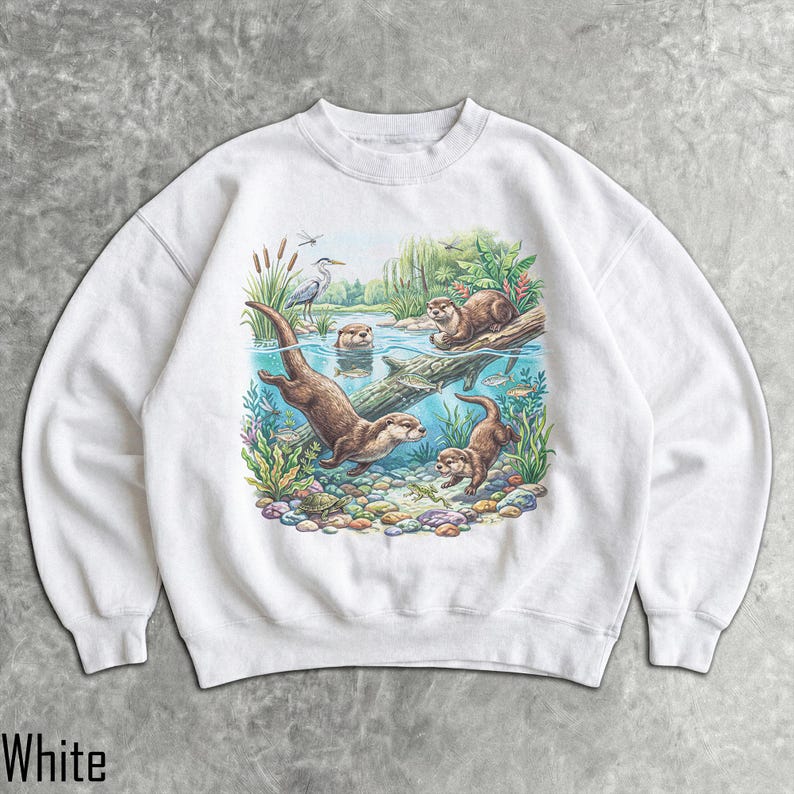 Op de afbeelding: Een wit sweatshirt met ronde hals en een kleurrijke grafische print van otters die in een rivier zwemmen. De illustratie toont meerdere otters, een reiger, waterplanten, vissen en een schildpad tussen kleurrijke rivierstenen.