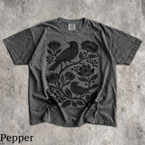 May include: A charcoal gray short-sleeve t-shirt featuring a black graphic print of two birds perched among stylized flowers and leaves. The shirt has a crew neckline and a faded, washed-out appearance with a visible brand tag at the inner collar.