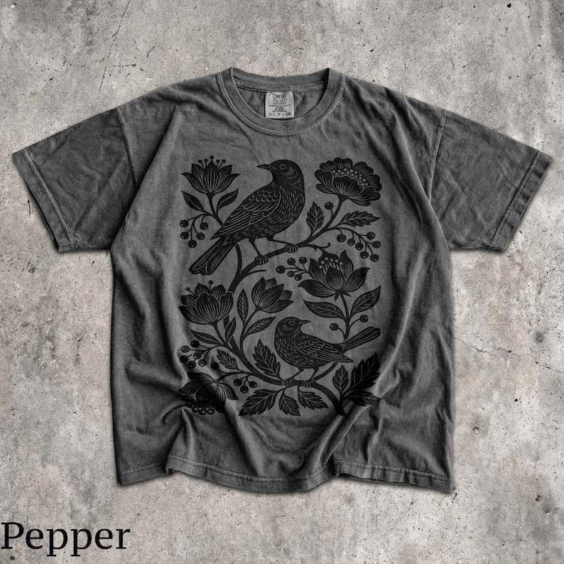 May include: A charcoal gray short-sleeve t-shirt featuring a black graphic print of two birds perched among stylized flowers and leaves. The shirt has a crew neckline and a faded, washed-out appearance with a visible brand tag at the inner collar.