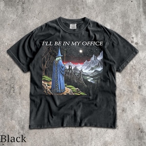 Op de afbeelding: Een zwart t-shirt met korte mouwen en een grafische print van een tovenaar in een blauwe mantel die op een klif staat en uitkijkt over een berglandschap bij nacht. De tekst I'LL BE IN MY OFFICE staat in het wit boven de illustratie.