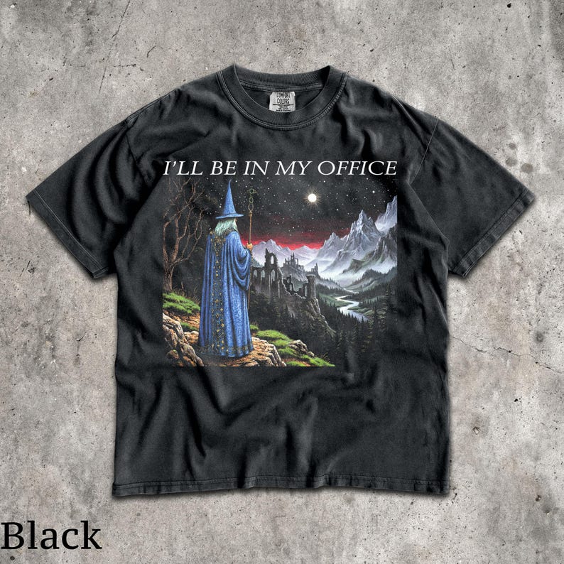 Op de afbeelding: Een zwart t-shirt met korte mouwen en een grafische print van een tovenaar in een blauwe mantel die op een klif staat en uitkijkt over een berglandschap bij nacht. De tekst I'LL BE IN MY OFFICE staat in het wit boven de illustratie.