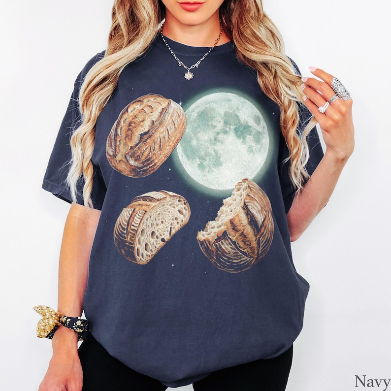 May include: A navy blue short-sleeve t-shirt featuring a graphic print of a large, glowing full moon centered above three distinct loaves of crusty sourdough bread. The bread loaves are arranged in a triangular formation around the moon.