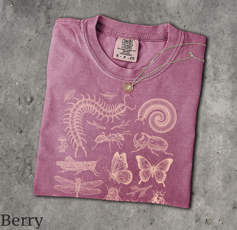 May include: A berry-colored short-sleeve t-shirt featuring a light-colored graphic print of various insects, including a centipede, millipede, beetle, grasshopper, butterflies, and a dragonfly. The shirt has a Comfort Colors brand label.
