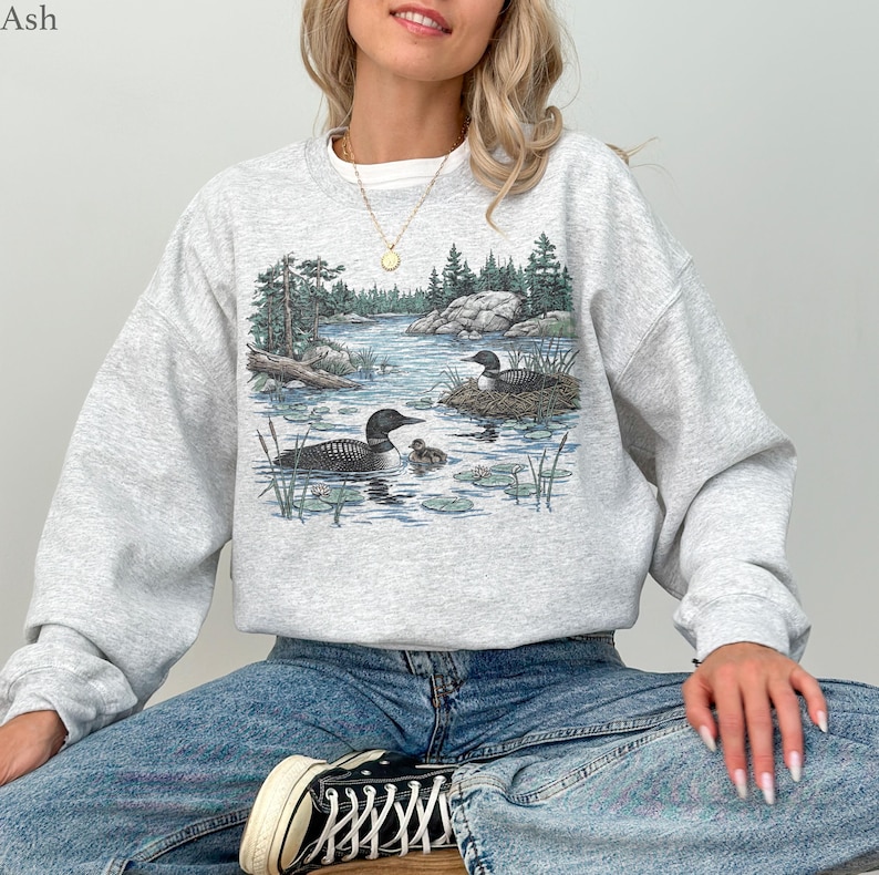 May include: A light gray crewneck sweatshirt featuring a detailed graphic print of two loons swimming in a lake with a small chick, surrounded by pine trees and rocky shores. The word Ash is printed in the top left corner of the image.