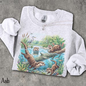 Op de afbeelding: Een asgrijs sweatshirt met ronde hals en een gedetailleerde grafische print van otters die in een rivier zwemmen. De illustratie bevat een reiger, waterplanten en vissen in een natuurlijke omgeving. Het kledingstuk heeft lange mouwen.