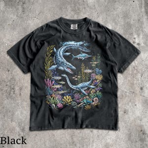 Op de afbeelding: Zwart t-shirt met een gedetailleerde illustratie van zeereptielen en vissen in een onderwaterlandschap. Het ontwerp bevat een grote mosasaurus en een plesiosaurus, omringd door kleurrijk koraal en zeewier. Het woord "Black" staat onderaan afgedrukt.