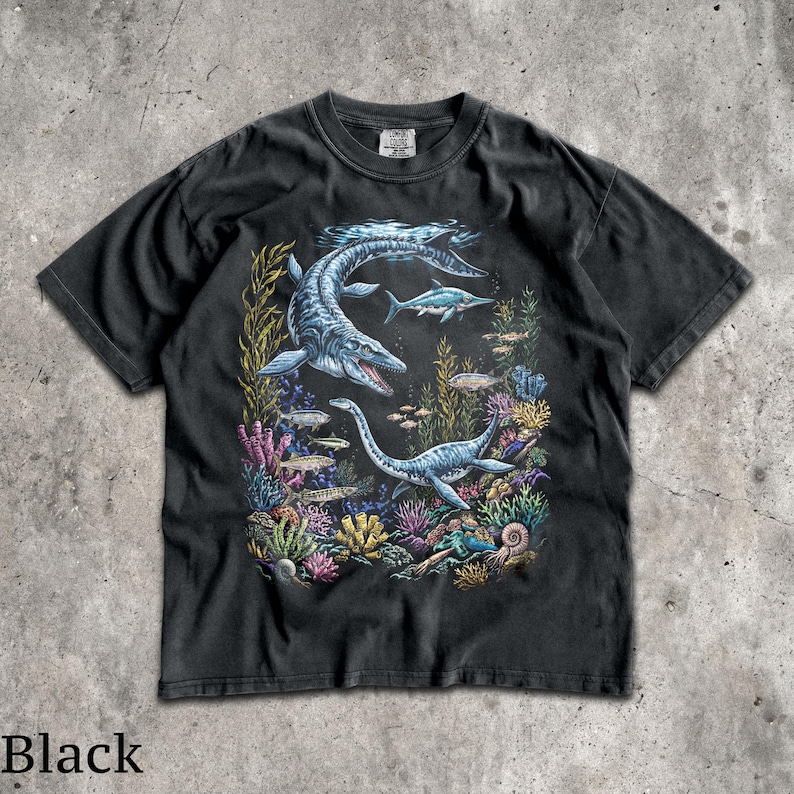 Op de afbeelding: Zwart t-shirt met een gedetailleerde illustratie van zeereptielen en vissen in een onderwaterlandschap. Het ontwerp bevat een grote mosasaurus en een plesiosaurus, omringd door kleurrijk koraal en zeewier. Het woord "Black" staat onderaan afgedrukt.