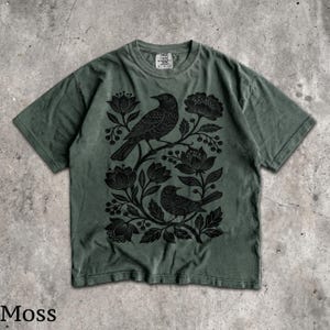 May include: A moss green short-sleeve t-shirt featuring a black graphic print of two birds perched among stylized flowers and berries. The shirt has a crew neckline and a tag at the inner collar that reads Comfort Colors with size S-P-CH.