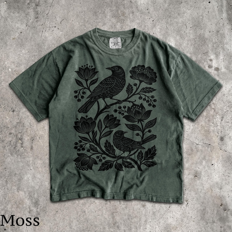 May include: A moss green short-sleeve t-shirt featuring a black graphic print of two birds perched among stylized flowers and berries. The shirt has a crew neckline and a tag at the inner collar that reads Comfort Colors with size S-P-CH.