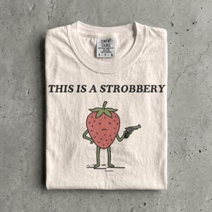 May include: Cream-colored t-shirt with the text "THIS IS A STROBBERY" above a cartoon strawberry holding a gun. The strawberry is red with green leaves and green legs. The shirt has a Comfort Colors tag.