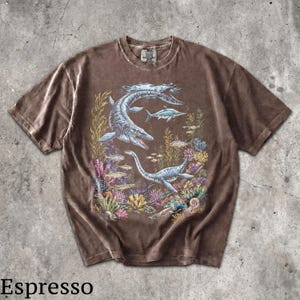 Op de afbeelding: Espresso-kleurig T-shirt met een gedetailleerde illustratie van prehistorisch zeeleven, waaronder plesiosauri&euml;rs en vissen, tegen een kleurrijke koraalrif achtergrond. Het shirt heeft een vintage, gewassen look.