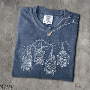 May include: A navy blue short-sleeve t-shirt featuring a white graphic print of four bats hanging from a tree branch against a starry night sky. The shirt has a crew neckline and a Comfort Colors brand label inside the collar with size S-CH.